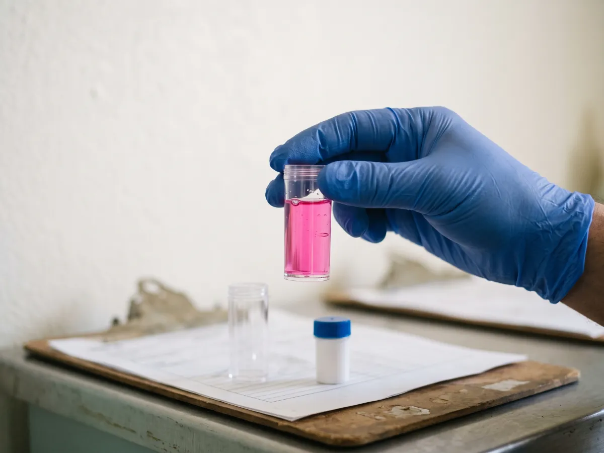 A gloved hand holding a comparator test cell of water turned pink by a chlorine test reagent, above a clipboard.