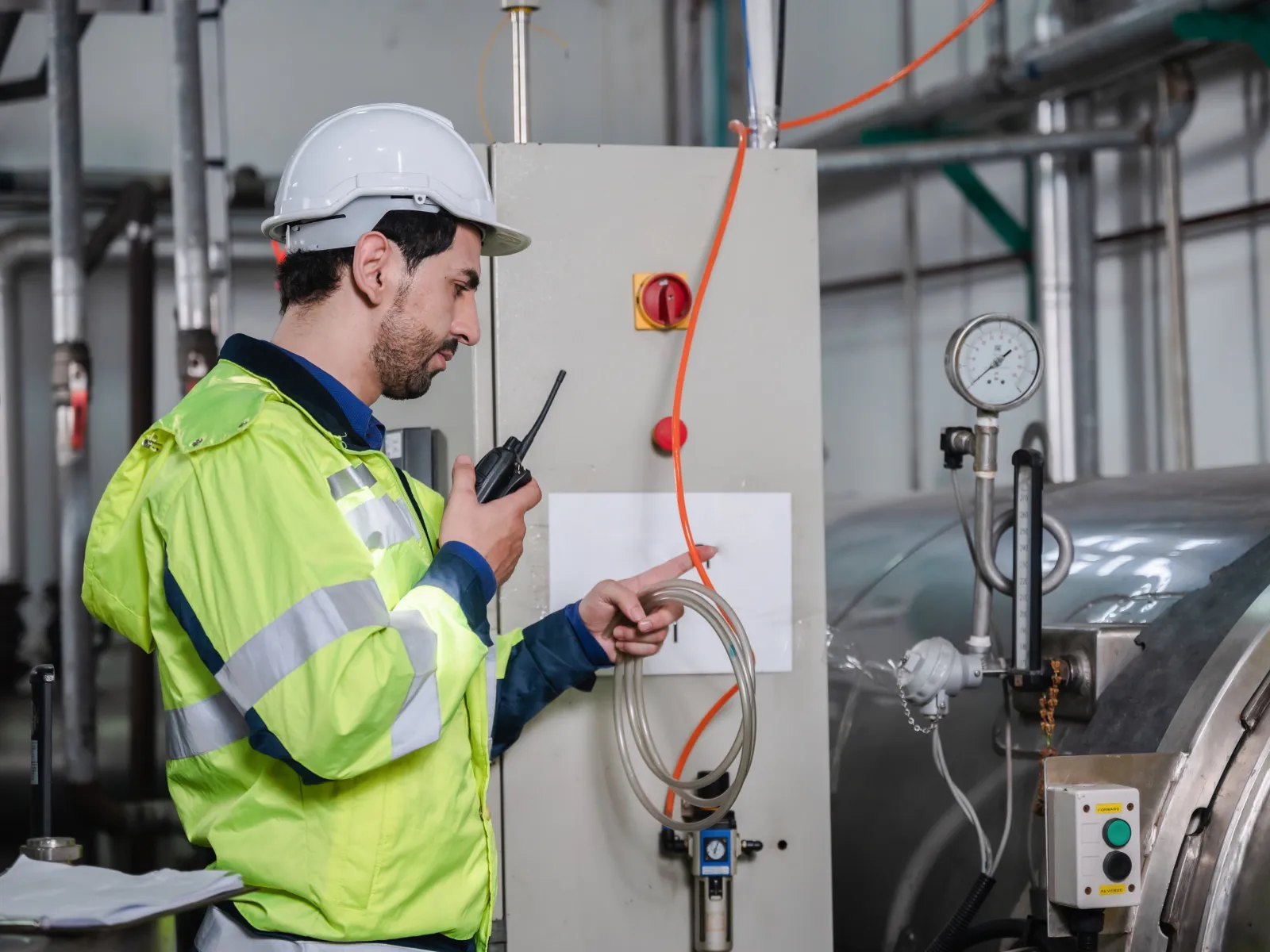 An engineer in a hard hat and hi-vis jacket using a two way radio at a control panel beside a large insulated water heating vessel.