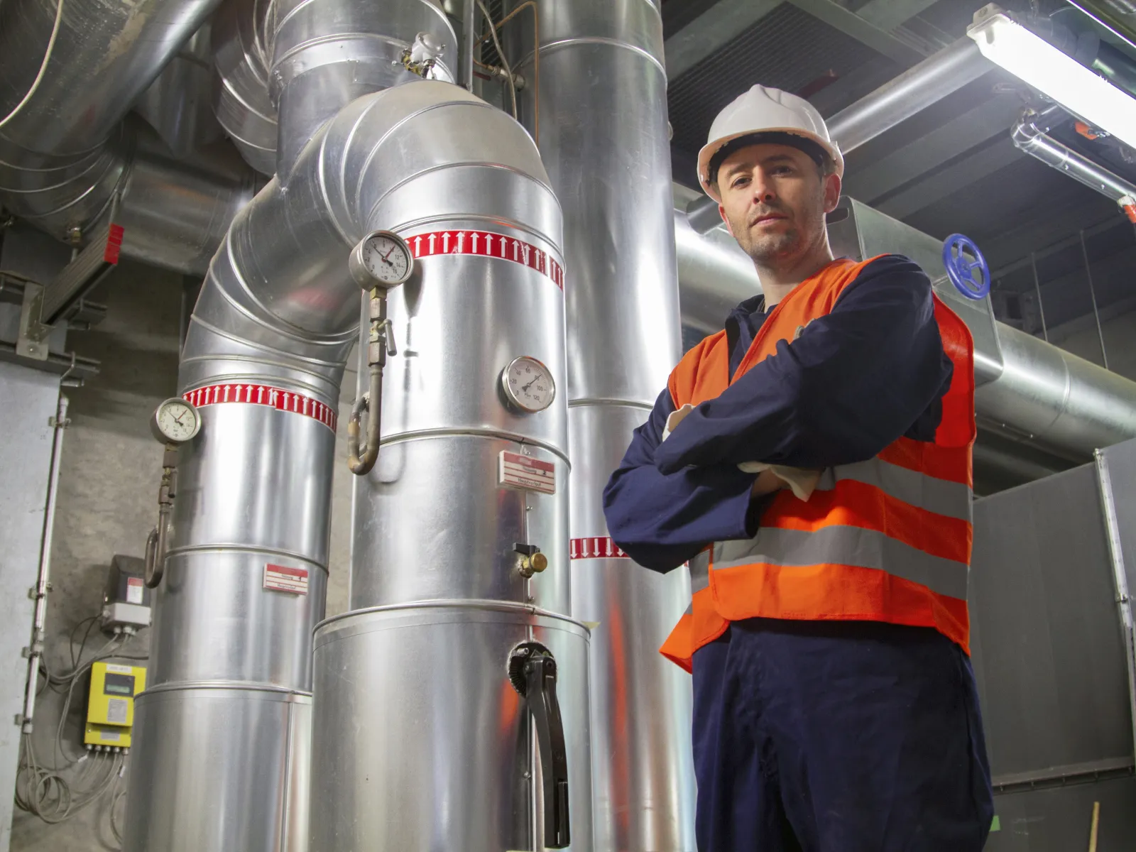 An engineer in a hard hat and hi-vis vest standing beside insulated stainless steel pipework and pressure vessels in a plant room.
